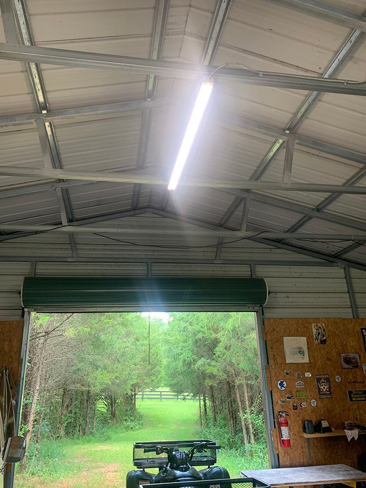 Interior of metal shed with open door revealing a tree-lined path. ATV parked inside. Fluorescent light on ceiling.