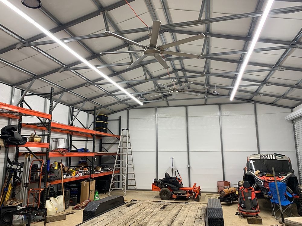 Interior of a large workshop with shelving, tools, and machinery; a large fan and LED lights hang from the ceiling.