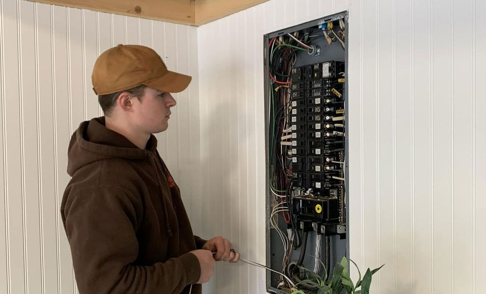 Man in brown hoodie and hat working on an electrical panel on a white wall.
