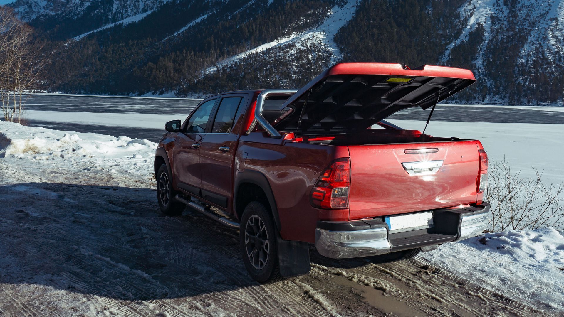 Red pickup truck with an open canopy on a snowy mountain road near a lake