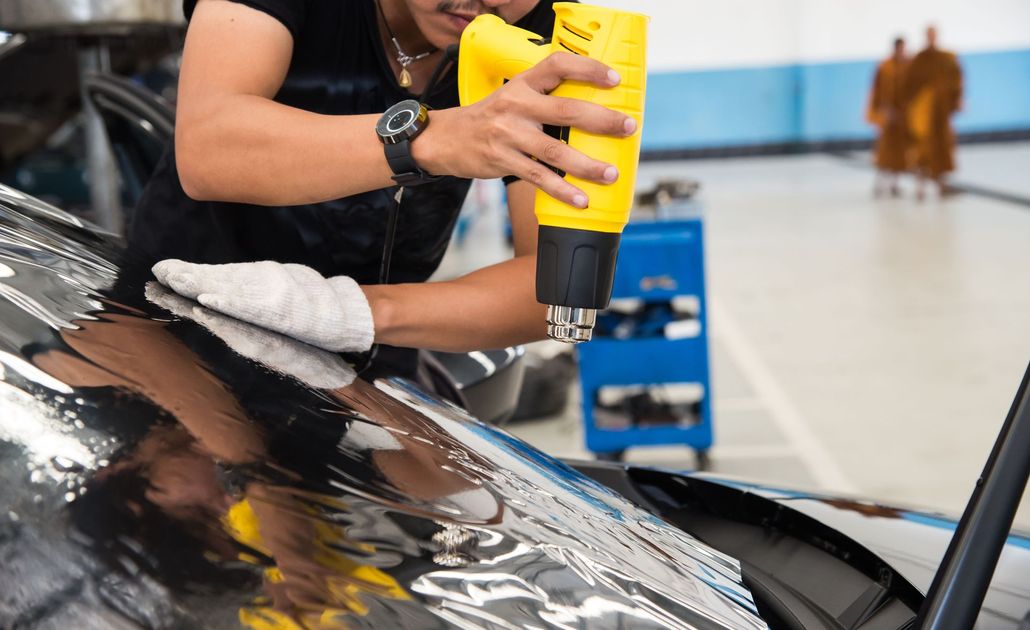 Person using a yellow heat gun to shrink-wrap film on a car hood in a garage