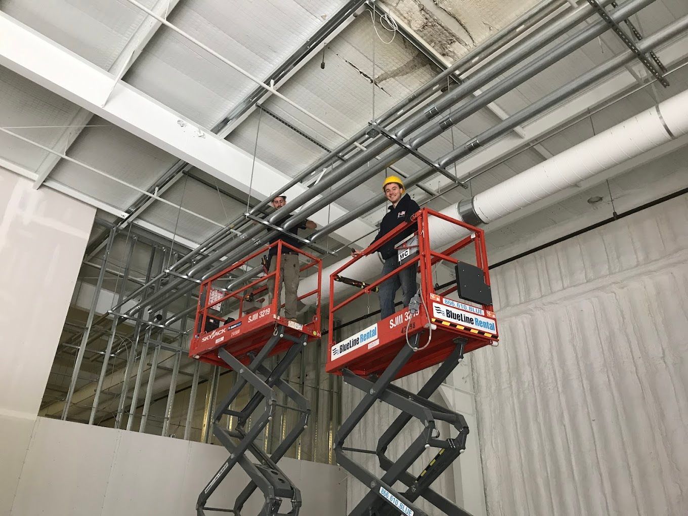Two workers on red scissor lifts inspecting pipes and ceiling panels in an industrial warehouse