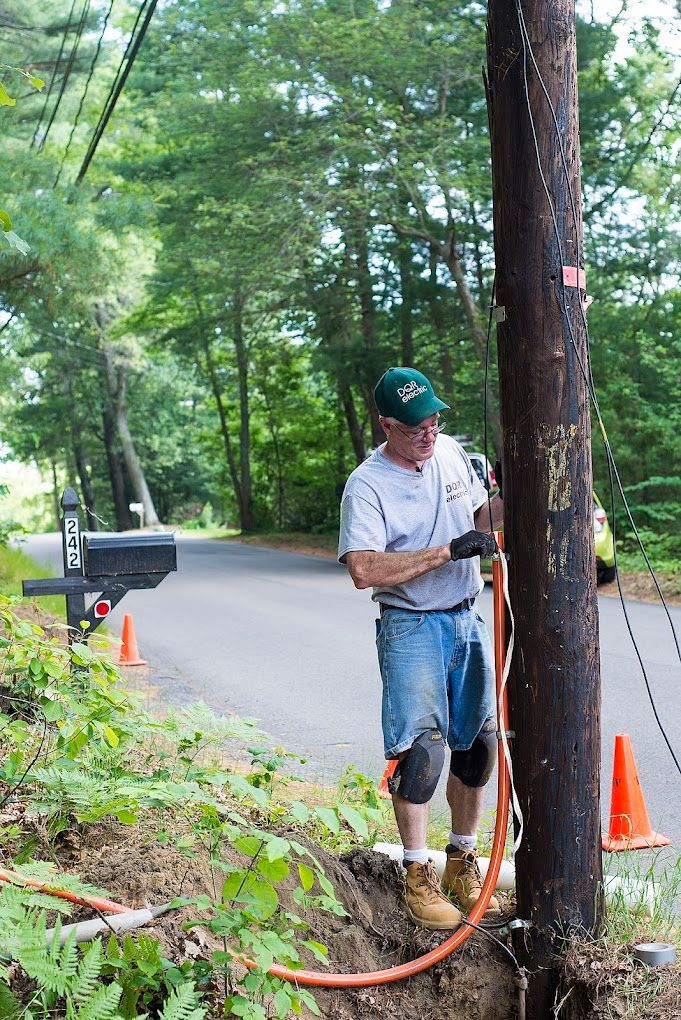 Worker in hard hat using a power tool on a roadside utility pole with orange cones nearby