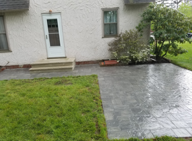 Gray brick patio next to green lawn and a house with a white door.