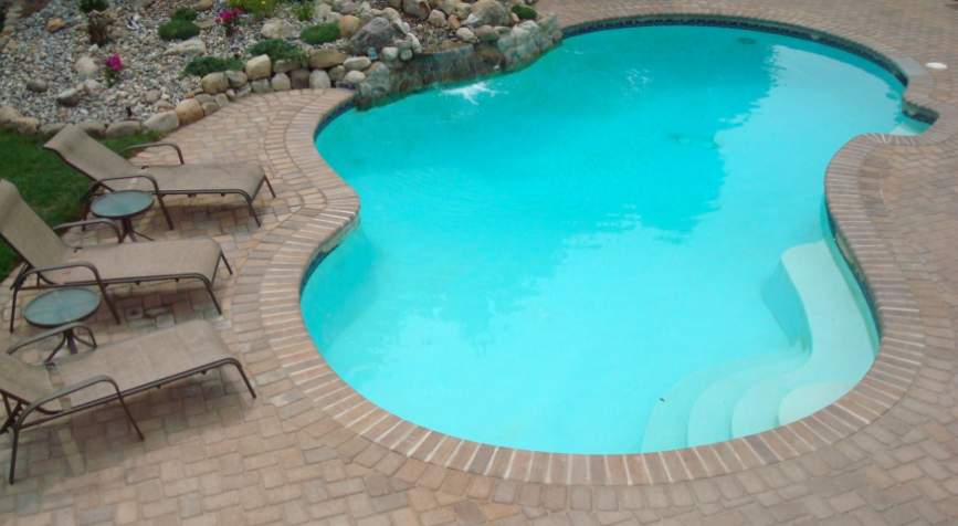 Pool with turquoise water surrounded by a brick patio, lounge chairs, and landscaping.