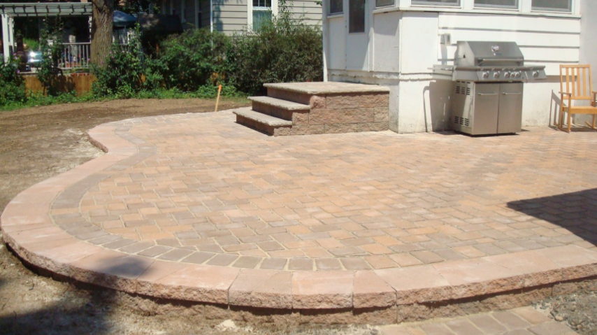 Brick patio with curved border, steps, and built-in grill near a house.