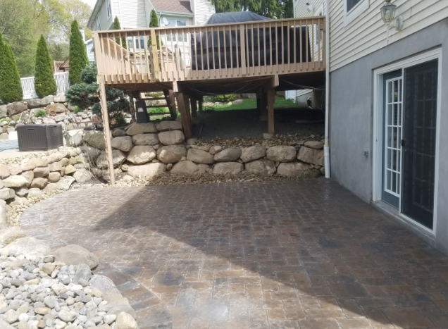 Stone retaining wall supports a wooden deck above a concrete patio, next to a house with a sliding glass door.