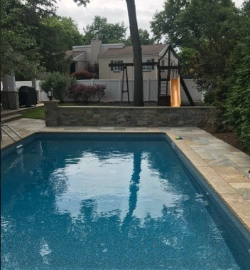 Pool with blue water, stone patio, brick wall, and a wooden playset in the background.