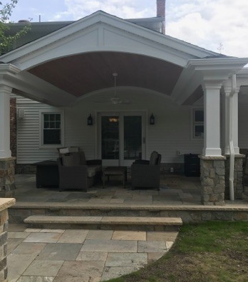 Covered patio with stone columns and seating, leading to a white house with a brick chimney.