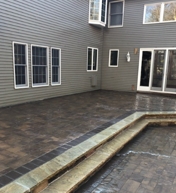Brick patio beside a house with gray siding and windows, with a low stone retaining wall.