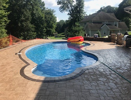 Swimming pool with blue water and stamped concrete patio, partially surrounded by trees and a house.