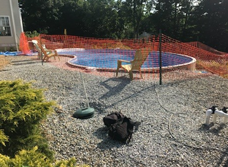 A nearly finished, kidney-shaped in-ground pool surrounded by gravel; chairs and orange safety fence nearby.