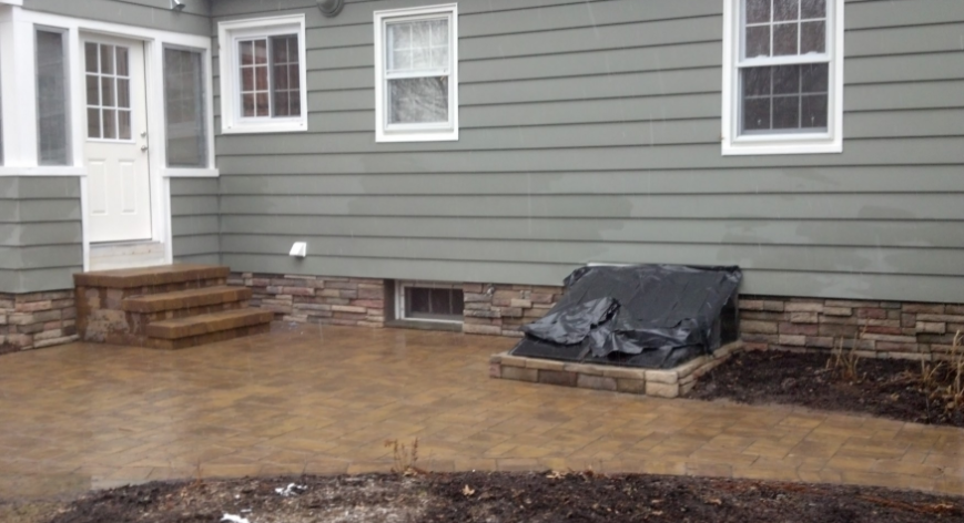 Exterior of house with gray siding, stone foundation, concrete patio, and covered window well.
