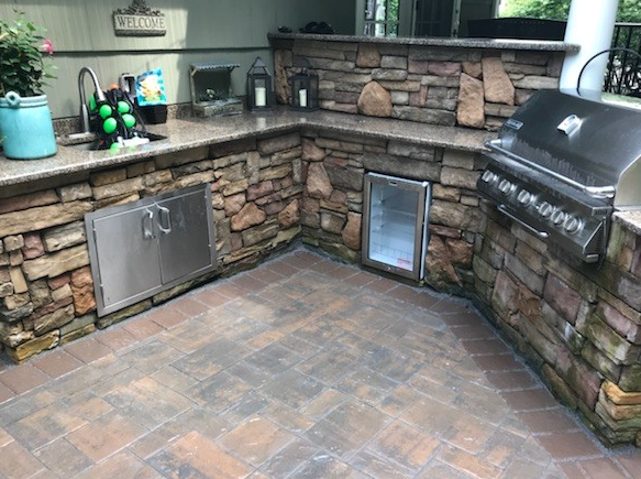 Outdoor kitchen with stone walls, stainless steel appliances, and a grill on a brick patio.