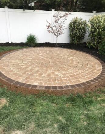 Circular brick patio with brown and tan pavers, surrounded by soil and green grass, set against a white fence.