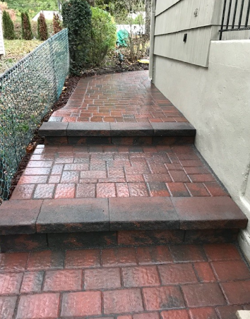 Brick paved steps leading to a building, with a chain-link fence on the left and landscaping in the background.