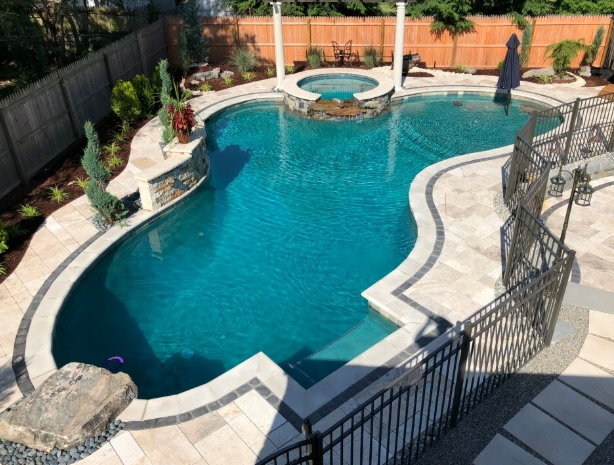 A large, teal-colored swimming pool with a hot tub, surrounded by stone patio, landscaping, and a wrought iron fence.