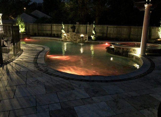 Pool at night, lit with colorful underwater lights. Paver patio with fence and home in the background.