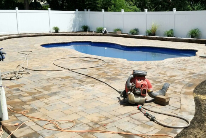 Backyard with a swimming pool and brick paving. A leaf blower is on the paving. White fence in the background.