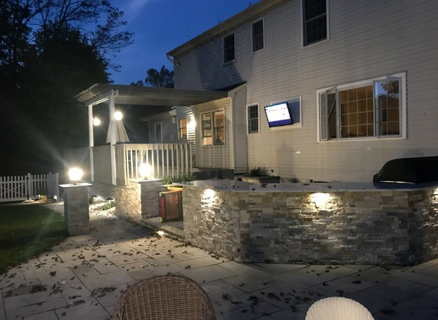 Backyard patio at dusk with a stone wall and lights, a house, deck, and TV.