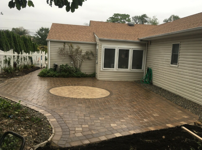 Brick patio with walkway, tan house with brown roof, white fence, and green hose.