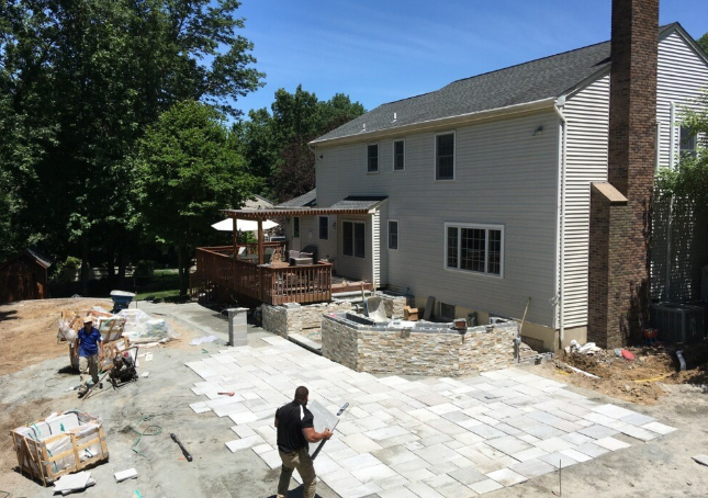 Workers installing a stone patio next to a two-story beige house with a wooden deck and chimney.