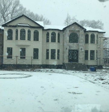 Two-story beige house with arched windows and a black door in a snowy setting. Scaffolding visible.
