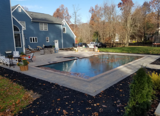 Backyard pool with paved patio next to a blue house, surrounded by grass and trees.