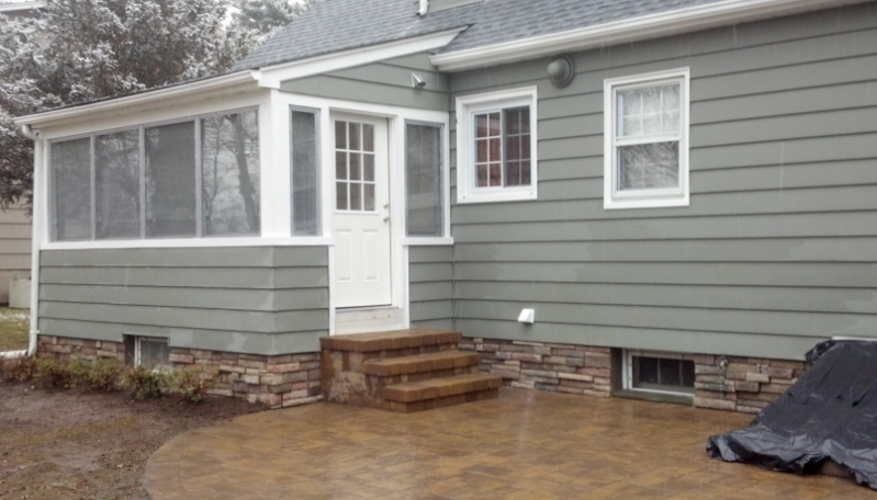 Back of a house with a stone foundation, a screened porch, and a patio. The siding is green.