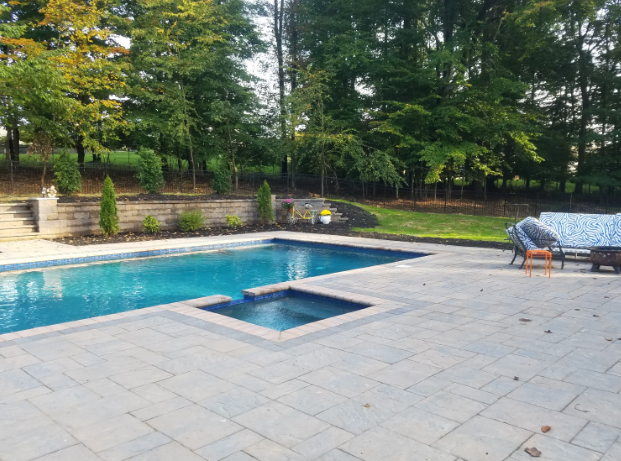 Pool with spa on a paved patio, overlooking a green lawn and trees.