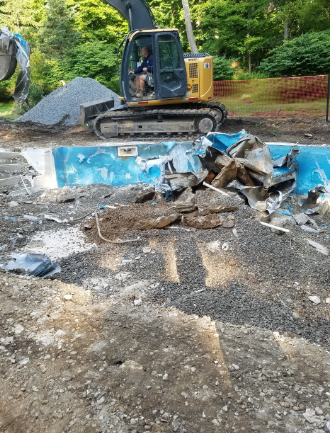 Excavator demolishing a swimming pool; operator inside. Blue pool interior, gravel, dirt, and trees in the background.