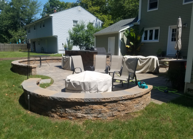 Stone patio with fire pit, seating, and built-in bar area in a backyard setting.
