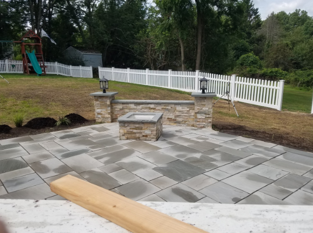 Patio with a stone fire pit, low wall, and decorative lights. White picket fence and a playground in the background.