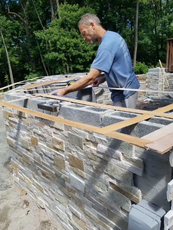 Man building an outdoor stone structure, using a level and other tools. Sunny day.