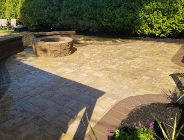 Brick patio with fire pit, bordering green shrubs and flower bed.