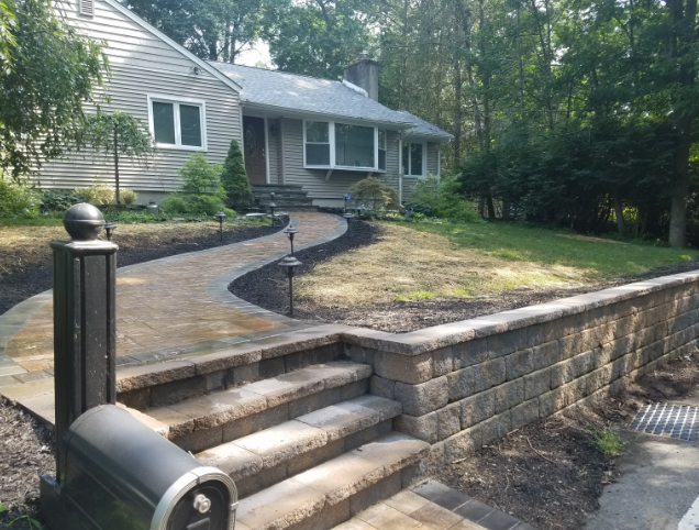 Stone walkway and steps leading to a gray house with a small lawn, and a dark mailbox post in the foreground.