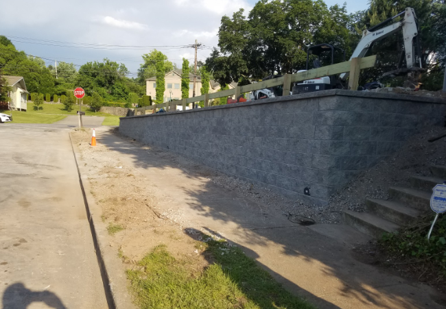 A large gray retaining wall borders a street, with a wooden fence on top. Excavator in the background.