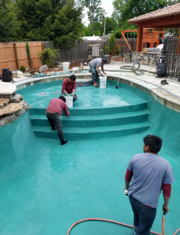 Workers applying paint to a teal-colored swimming pool. One holds a bucket, another brushes the steps. Outdoor setting.