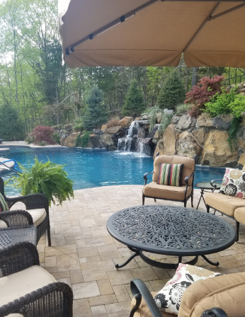 Patio with seating overlooks a pool with waterfall feature, set in a natural, wooded environment.