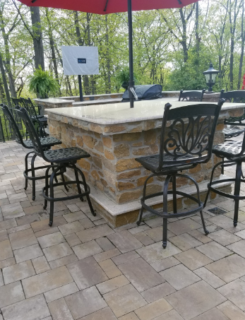Outdoor patio with stone bar, black chairs, and red umbrella; trees in background.