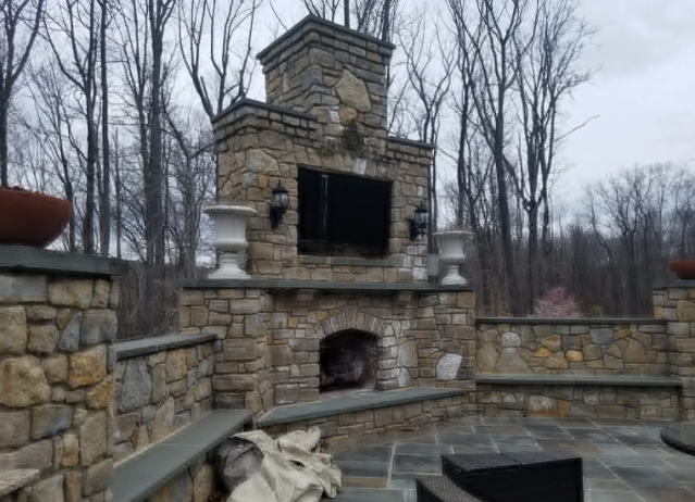 Stone outdoor fireplace with built-in TV, flanked by sconces and urns, set on a stone patio in wooded area.
