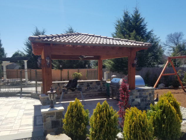 Outdoor kitchen with tiled roof, stone columns, and grill, set on a patio, under a clear blue sky.