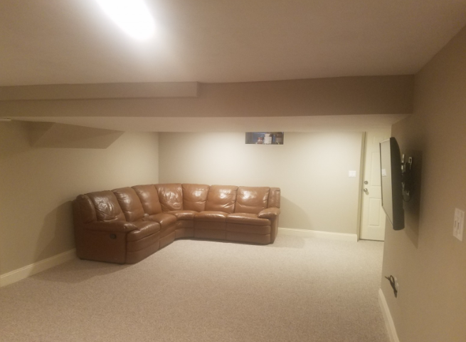 A basement room with a brown sectional sofa, TV, and door, beige walls and carpet.