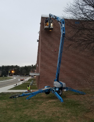 A blue articulated boom lift reaches up to a brick building; a person works in the lift's bucket.