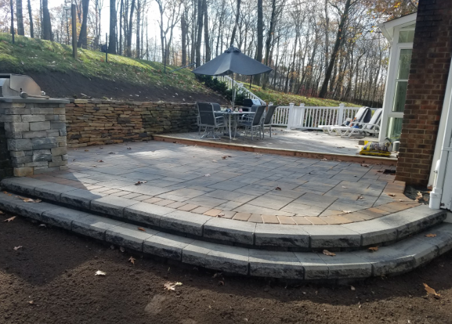 Stone patio with steps and retaining wall. Dining set on patio with a sloped hillside in the background.