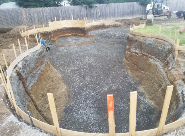 Pool construction site: Excavated earth, wooden forms, gravel base. Person working, machinery in background.
