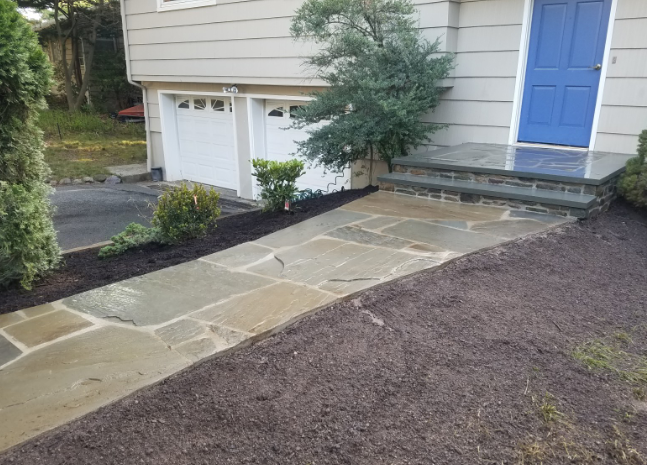 Stone walkway leading to a blue door and garage, lined with dark mulch and greenery.