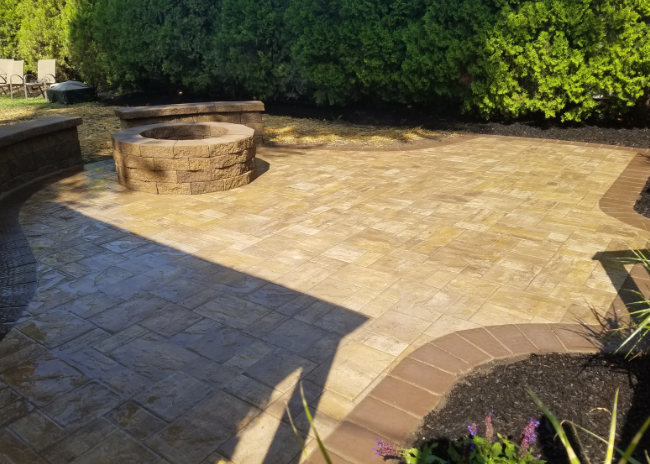 Brick patio with fire pit and dark brick border. Green bushes in background.