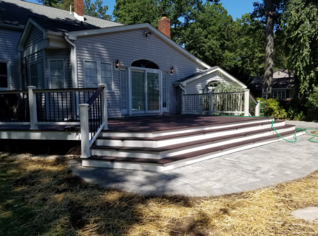 Backyard deck with multi-level stairs leading to a patio beside a house; brown deck boards, white railings.