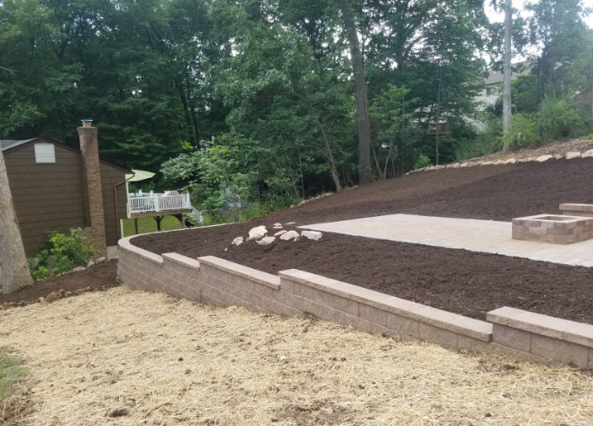 Terraced backyard with retaining walls, a patio, fire pit, and shed against a backdrop of trees.